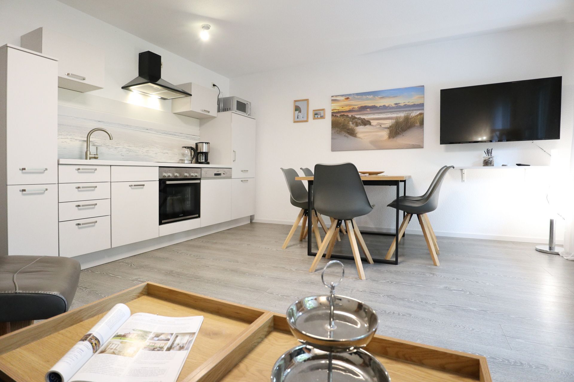 A modern kitchen with white cabinetry, a built-in oven, and a microwave. There is a dining area with a table and chairs, and a flat-screen TV mounted on the wall.