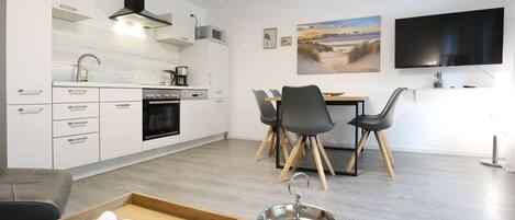 A modern kitchen with white cabinetry, a built-in oven, and a microwave. There is a dining area with a table and chairs, and a flat-screen TV mounted on the wall.