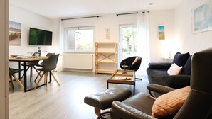 Ein modernes Wohnzimmer mit einer Couch, einem Esstisch mit Stühlen, einem an der Wand befestigten Fernseher und einem Fenster mit Blick auf Bäume.