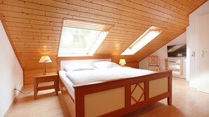 Ein Schlafzimmer mit schrägverdeckter Decke, Holzbalken, einem Bett mit weißer Bettwäsche, zwei Nachttischlampen und einem an der Wand montierten Fernseher.
