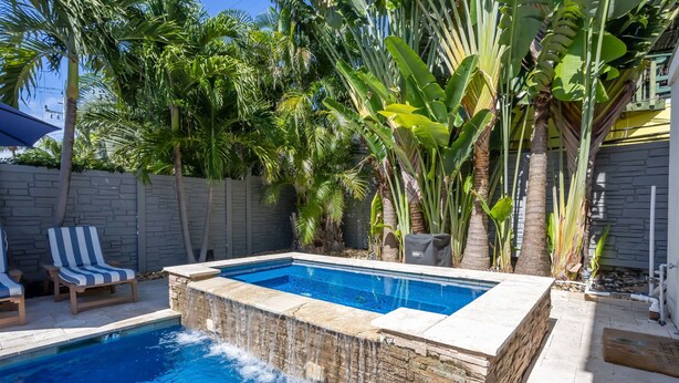 A swimming pool with a waterfall feature, surrounded by palm trees and a stone wall.