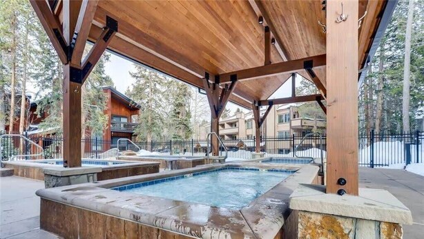 A wooden outdoor hot tub area with stone walls and a covered roof, surrounded by a fence and buildings in the background. - Timbernest Condo - 2 BDR Walk to Downtown & Lift! (Breckenridge)