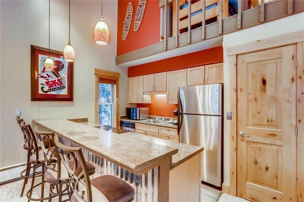 Una cocina con isla central, armarios de madera, refrigerador de acero inoxidable y luces colgantes.