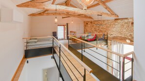 A spacious loft with exposed wooden beams, a stone wall, and a mezzanine level with a staircase.