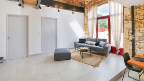 A spacious living area with a grey sofa, ottoman, and coffee table. There are two doors, a stone wall, and a view of greenery outside.