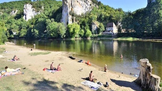 A riverside beach with people sunbathing and swimming, surrounded by greenery and a large rock formation.
