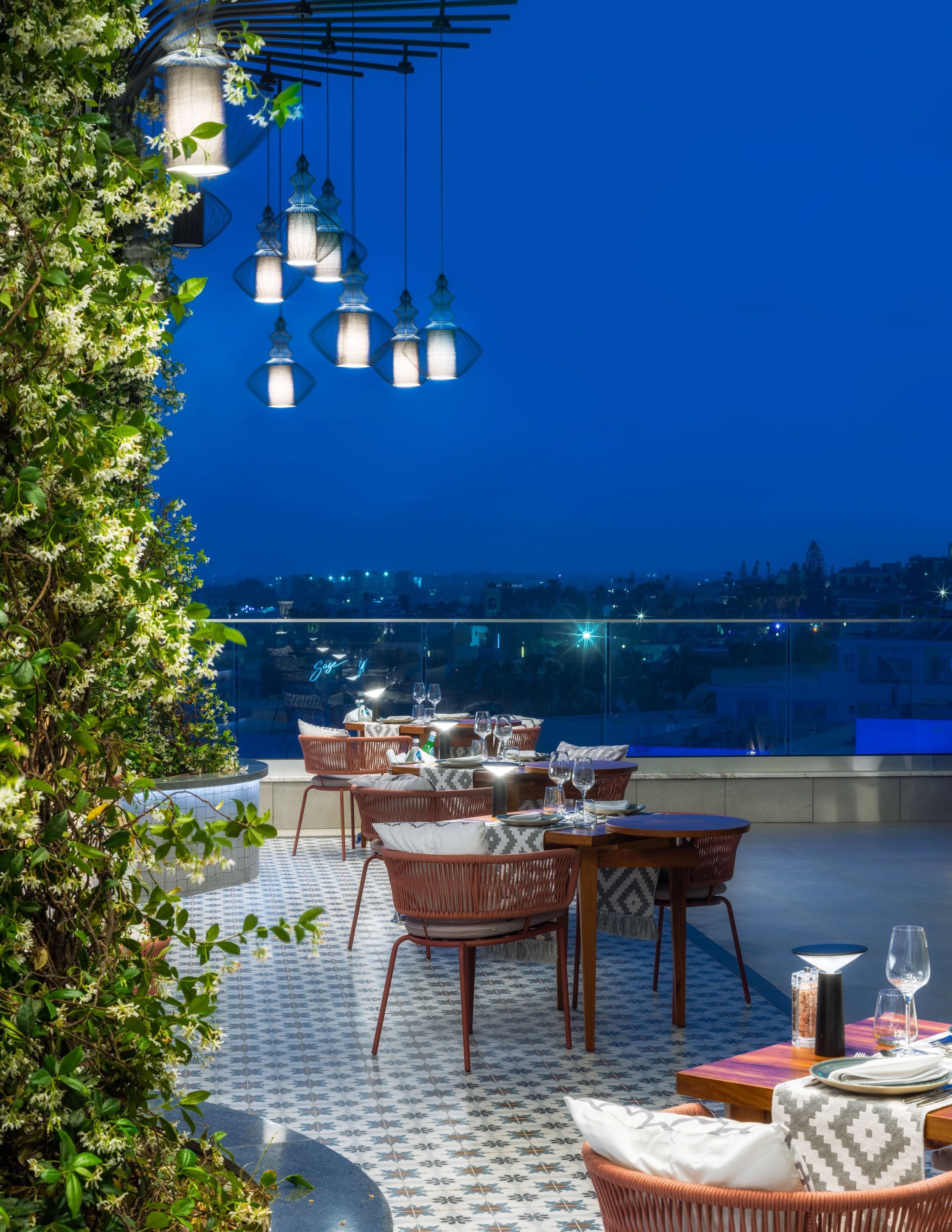 Un área de comedor al aire libre con mobiliario de mimbre y luces colgantes, con vistas a un paisaje urbano al atardecer.