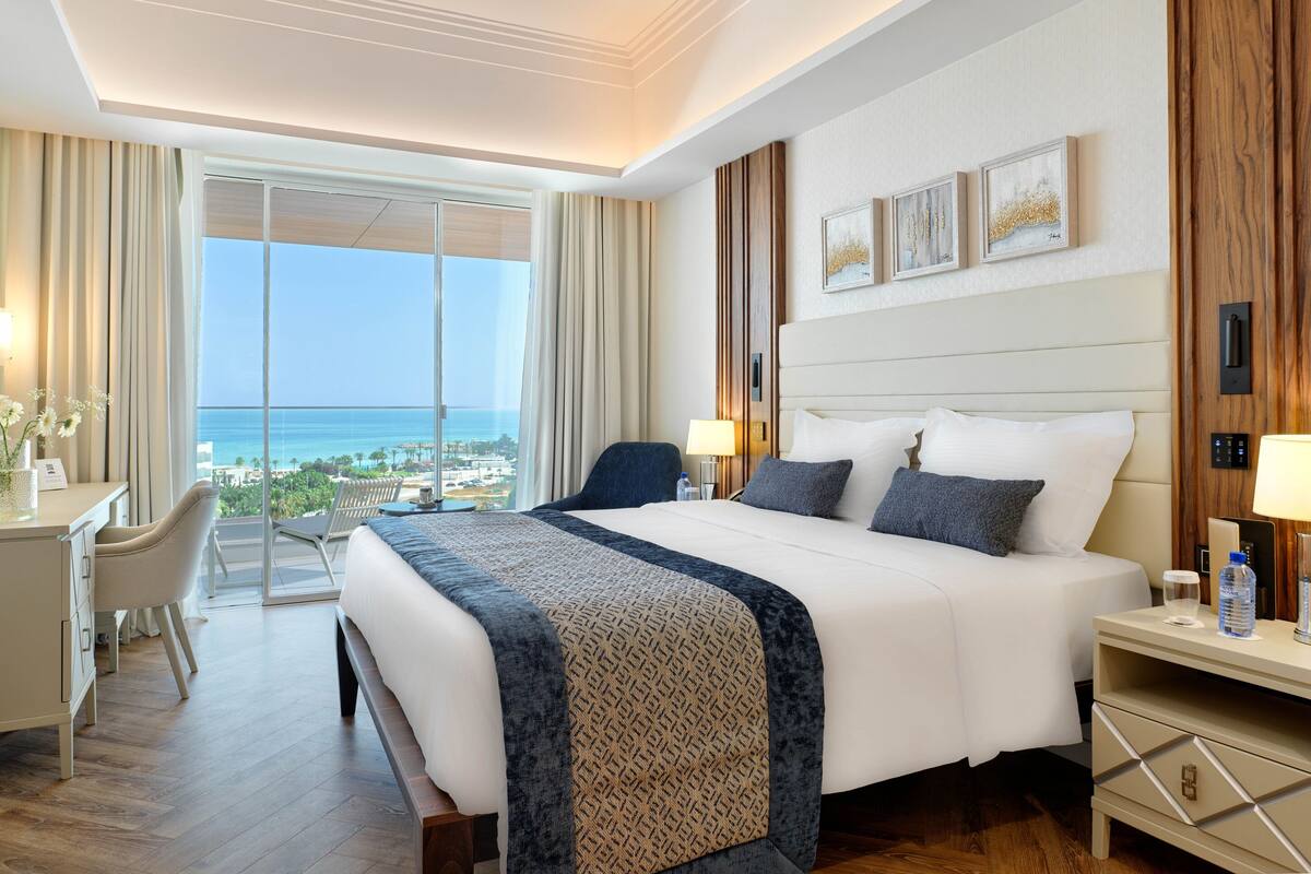 A hotel room with a large bed, bedside tables, a desk, and a view of the ocean.