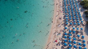 An aerial view of a beach with clear turquoise waters, numerous blue umbrellas, and people swimming and sunbathing. - Amanti, MadeForTwo Hotels – Ayia Napa (Ayia Napa)