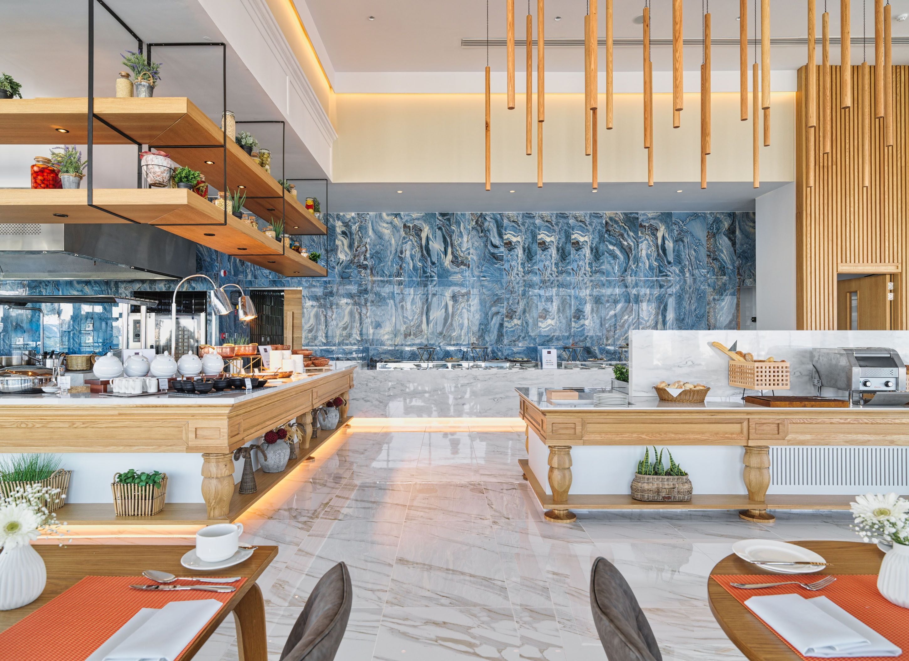 A modern restaurant with a marble backsplash, wooden shelves with plants, hanging wooden elements, and a central dining area with tables and chairs.