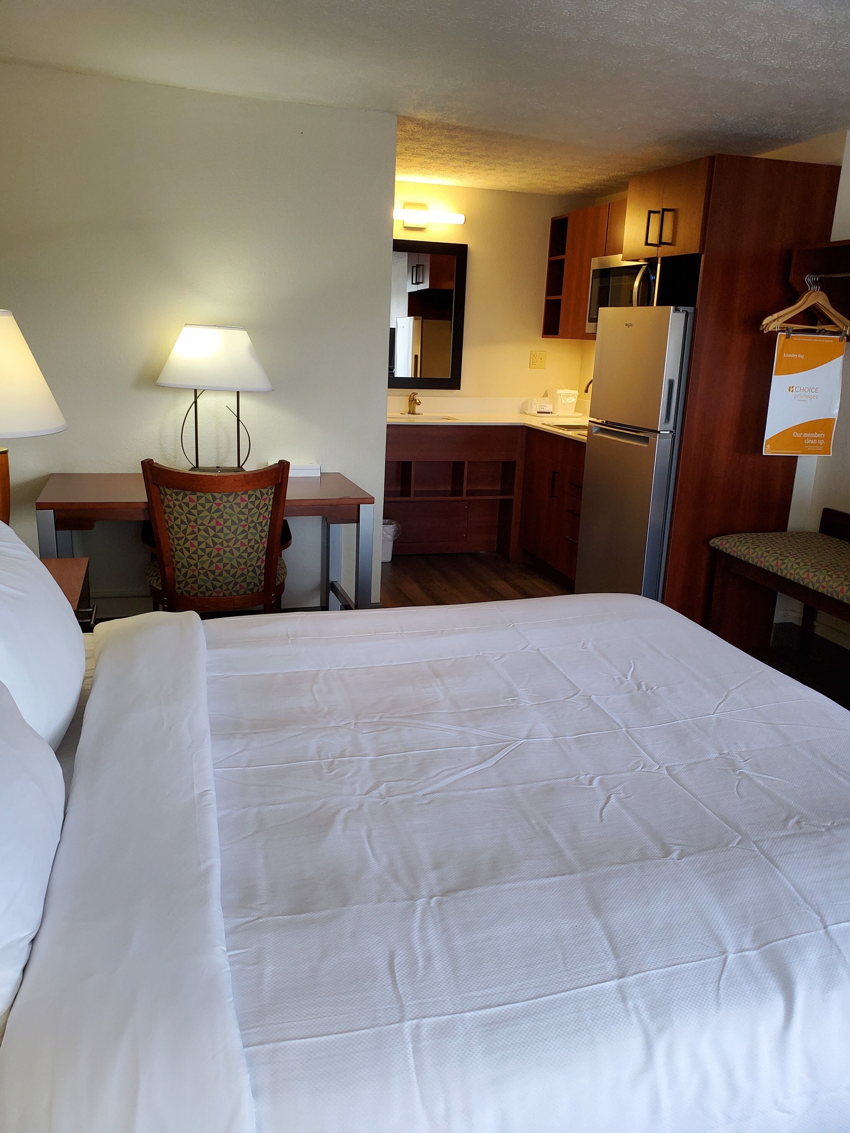 Una habitación de hotel con una cama grande, un escritorio con lámpara, una silla, una kitchenette con refrigerador y una bolsa para ropa sucia.