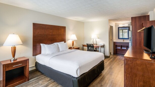 A hotel room with a large bed, two bedside tables with lamps, a desk with a chair, a television, and a bathroom visible through an open door. - Suburban Studios Middlesboro (Middlesboro)