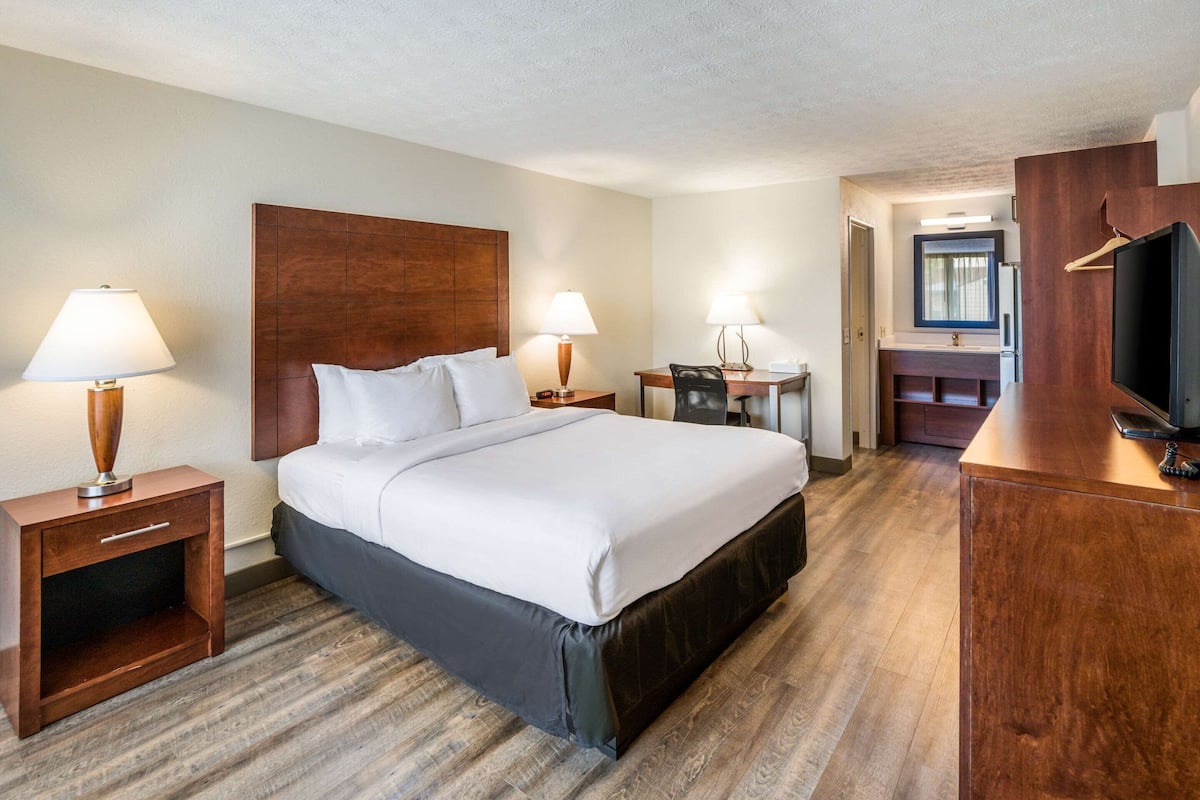 A hotel room with a large bed, two bedside tables with lamps, a desk with a chair, a television, and a bathroom visible through an open door.