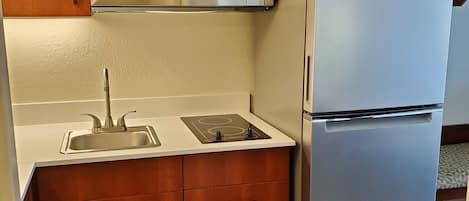 Una cocina compacta con armarios de madera, un microondas de acero inoxidable, horno integrado, un lavamanos y un refrigerador.
