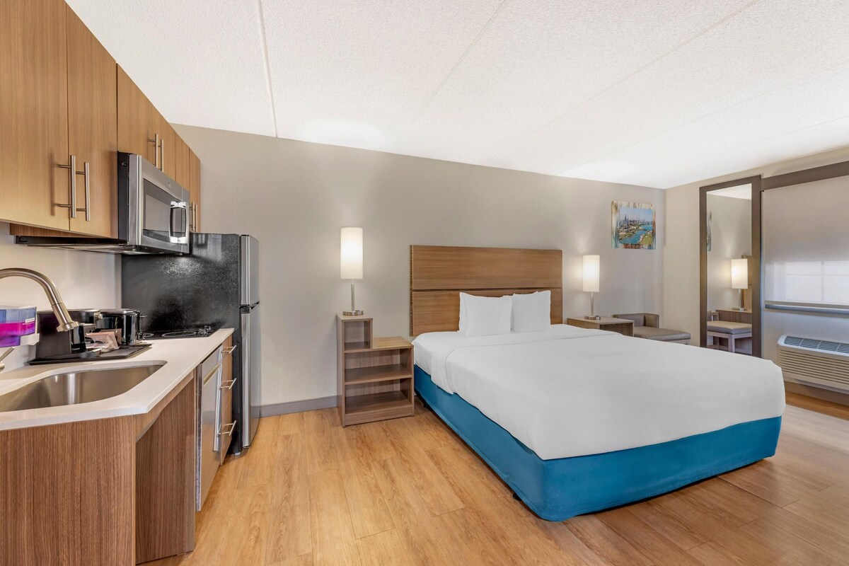 A modern hotel room with a kitchenette, a bed with a blue accent, and a large window.