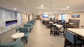 A spacious hotel lobby with a long row of blue chairs and tables, a flat-screen TV mounted on the wall, and a cityscape painting on the wall. - MainStay Suites Oak Brook Terrace-Chicago (Villa Park)