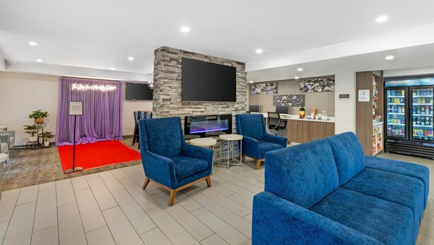 A modern lounge area with blue upholstered chairs, a flat-screen TV, a fireplace, and a bar area with a refrigerator stocked with beverages. - MainStay Suites Oak Brook Terrace-Chicago (Villa Park)