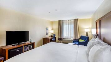 A hotel room with a large bed, a television, a desk with a computer, a chair, and an air conditioning unit.