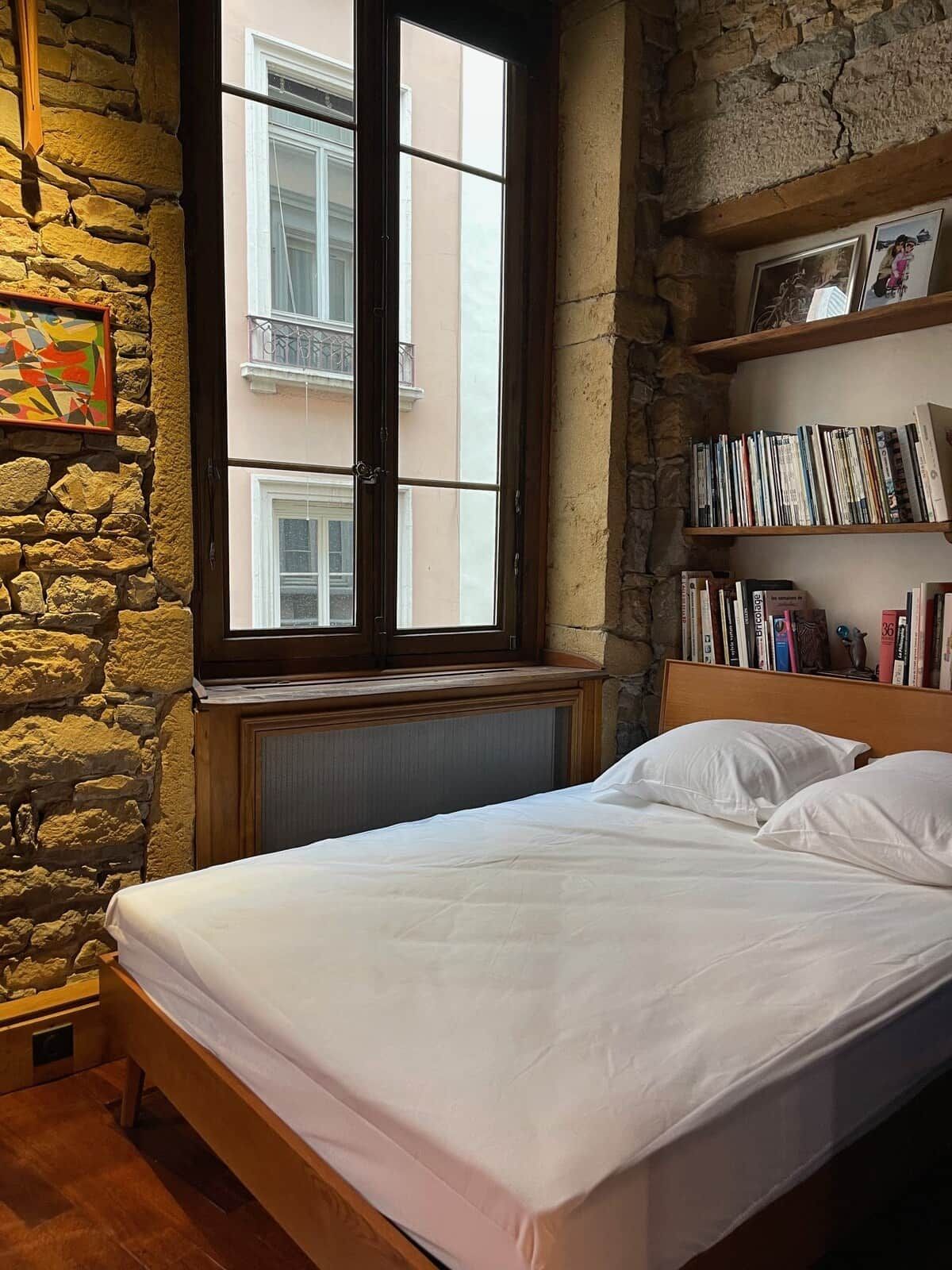 A bedroom with a stone wall, a window with a view of buildings, a bookshelf with books, and a bed with white bedding.