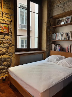 A bedroom with a stone wall, a window with a view of buildings, a bookshelf with books, and a bed with white bedding. - Charming "Heart of Lyon" Apartment with Elevator, A/C & Parking (Lyon)