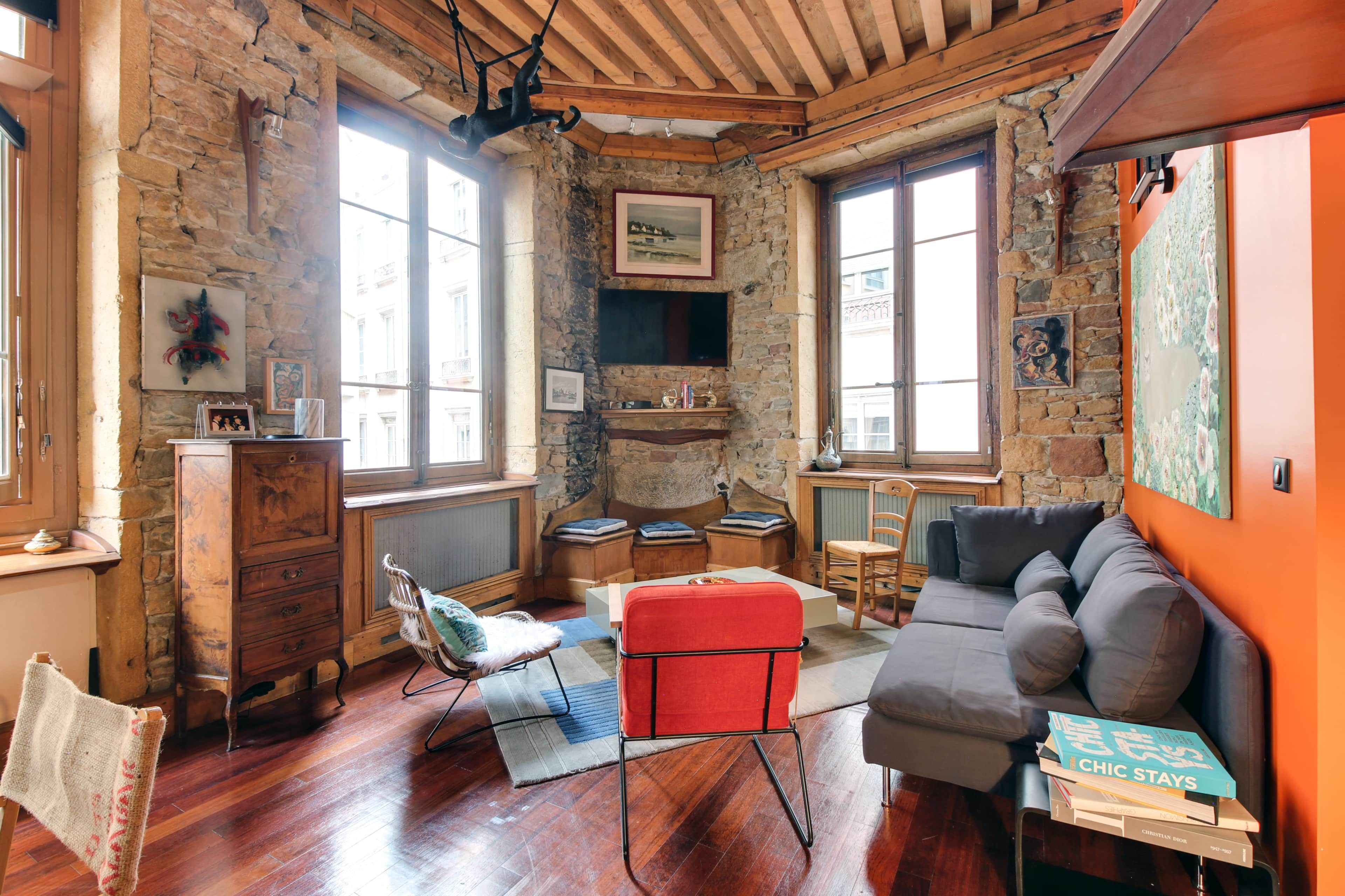 Ein gemütliches Zimmer mit einer Steinwand, Holzbalkendecke und einem roten Sessel. Auf einem kleinen Tisch liegen Bücher und ein Sofa mit Kissen.