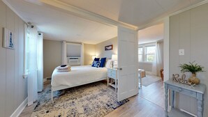 Une chambre à coucher comprenant un lit, des tables de chevet, une commode, une fenêtre avec des rideaux, un climatiseur fixé au mur et une porte donnant sur une autre pièce.