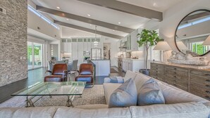 A spacious living area with a glass coffee table, leather chairs, and a sofa. A kitchen is visible in the background with white cabinets and a large island.