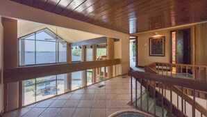 A spacious interior with a high ceiling, large windows offering a view of the sea, and a wooden staircase leading to an upper level.
