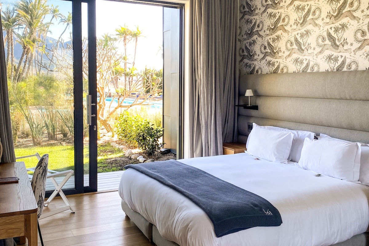 Ein Hotelzimmer mit einem großen Bett, einem Schreibtisch und Blick auf Pool und Grünflächen.