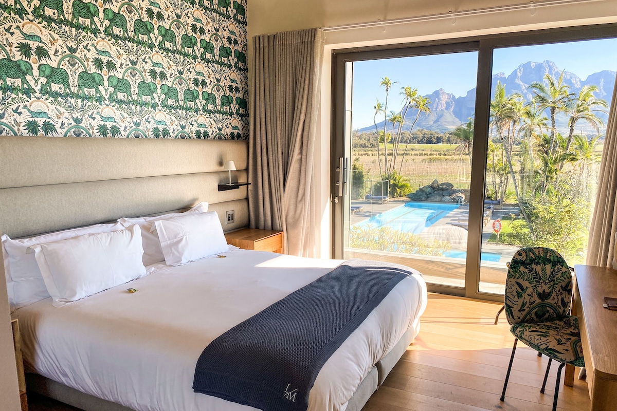 Ein Hotelzimmer mit einem großen Bett, Blick auf einen Pool und Berge sowie einer Wand mit Muster.
