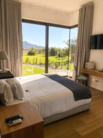 A bedroom with a large bed, a desk, a chair, a TV, and a view of a green landscape.