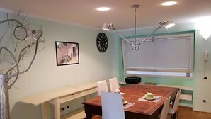 A dining area with a wooden table, white chairs, a clock, and a window with blinds. - Ambra - Central Apartment in Bergamo (Bergamo)