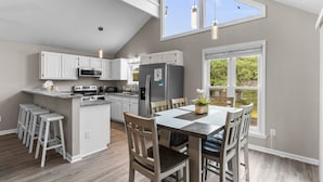 A modern kitchen with white cabinets, a central island with stools, and a dining area with a table and chairs. - Two Decks, Wheelchair Accessible, Large Yard with close beach access.  Sail Away (Oak Island)