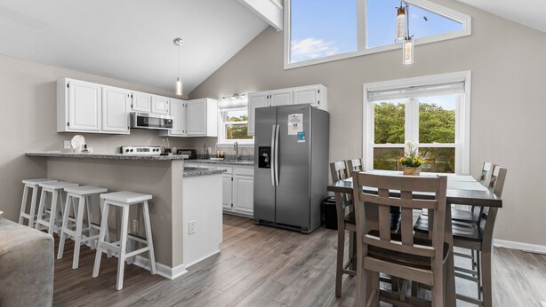 A modern kitchen with white cabinets, a stainless steel refrigerator, and a dining area with wooden chairs and a table. - Two Decks, Wheelchair Accessible, Large Yard with close beach access.  Sail Away (Oak Island)