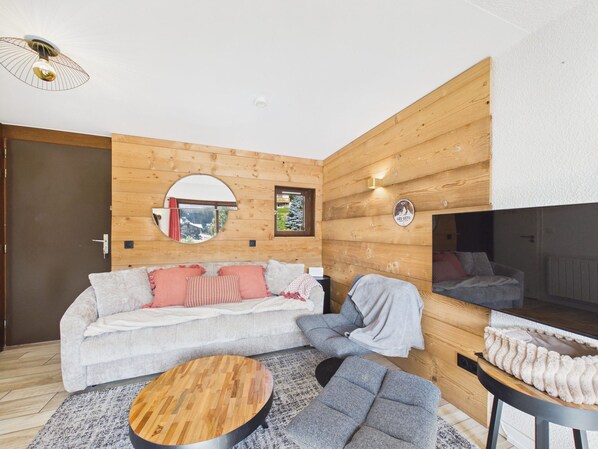 A modern living room with a wooden wall panel, a round mirror, a sofa with cushions, a coffee table, and a TV.