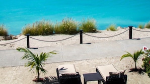 Une plage à l’eau turquoise et limpide, une zone sablonneuse avec des chaises longues, et une vue sur des montagnes lointaines.