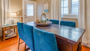 A dining area with a wooden table and blue upholstered chairs, a small side table with a lamp, and a window with white curtains. - Perfect Couples Getaway, Heated Pool Access (Tybee Island)