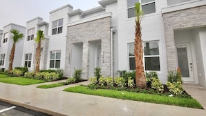A row of modern, white buildings with stone accents, featuring uniform windows and doors, and well-maintained landscaping with palm trees and shrubs. - Amazing NEW 3 bedroom+loft townhouse in Hidden Forest, with private pool! (Clermont)