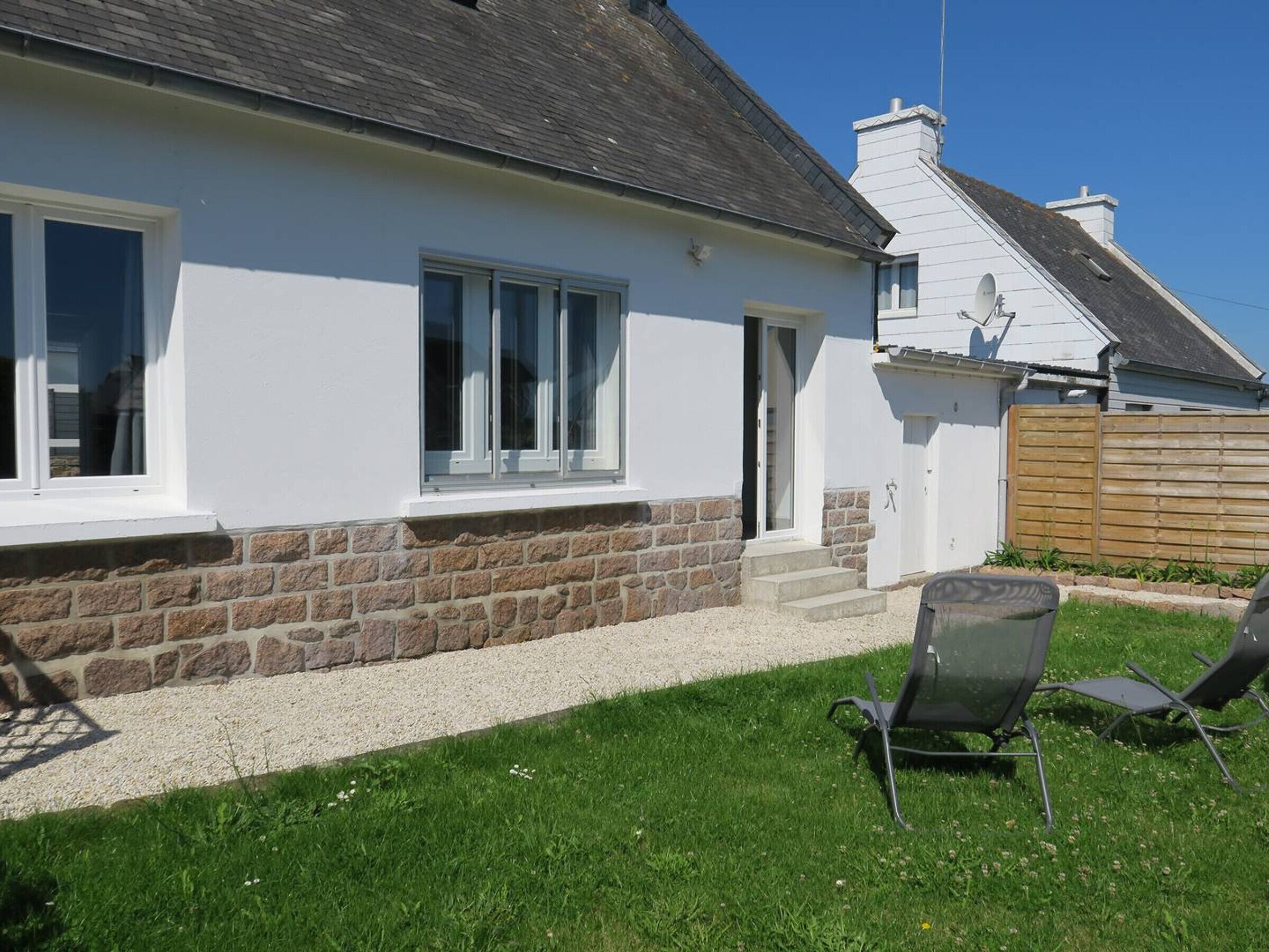 Une maison blanche avec des fondations en pierre, une porte d’entrée et un petit jardin devant, avec une pelouse et deux chaises.