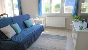 Ein modernes Wohnzimmer mit einem blauen Sofa, einem kleinen weißen Tisch, einem Heizkörper und einem Fernseher auf einem weißen Standfuß.
