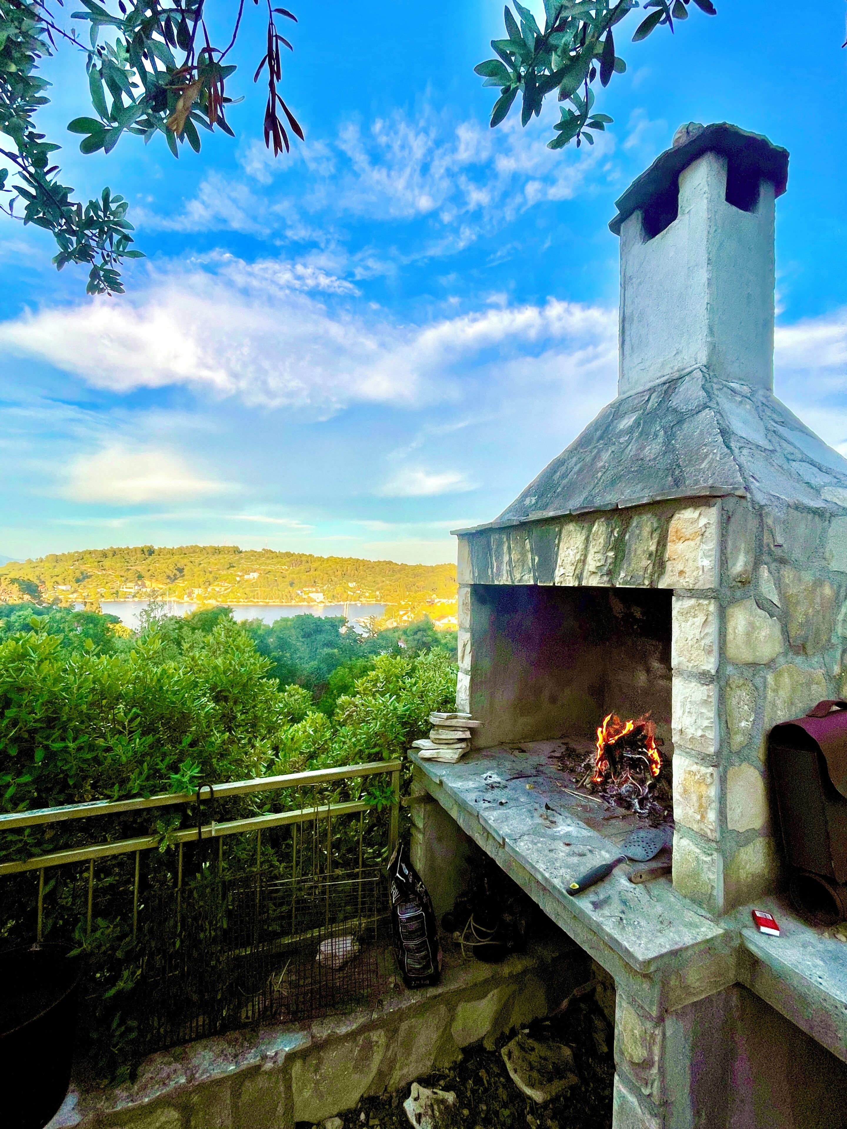 A stone-built outdoor oven with a fire burning inside, surrounded by greenery and a clear sky.