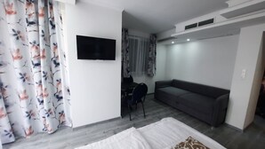 Une chambre d’hôtel moderne, équipée d’un lit, d’un canapé gris, d’un bureau avec une chaise et d’un téléviseur fixé au mur.