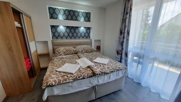 Une chambre moderne avec un grand lit, une armoire, une tête de lit dotée d’un éclairage intégré et une vue dégagée à travers des rideaux légers.