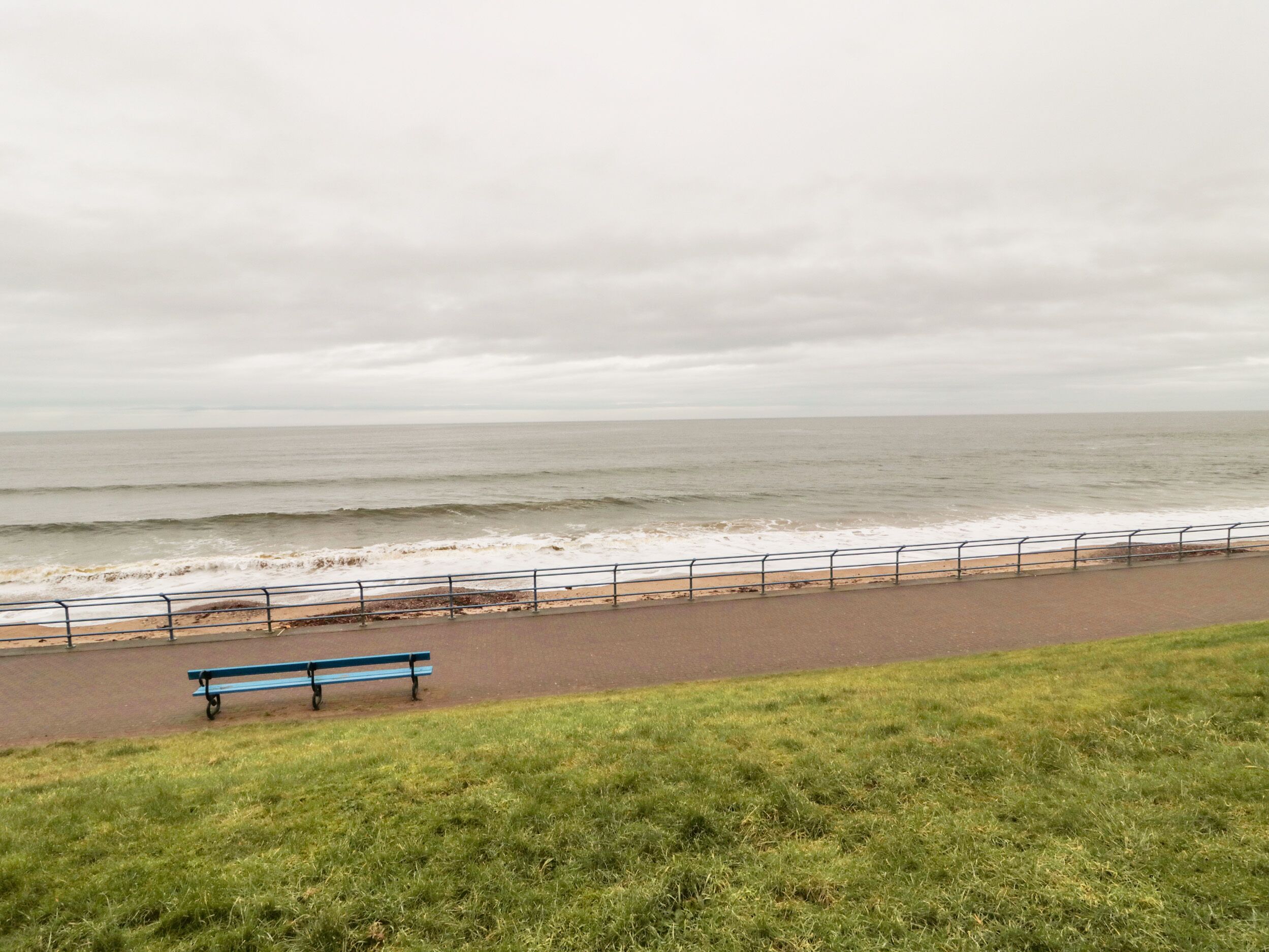 Un banc placé sur un chemin pavé, au bord d’une plage protégée par une rambarde, sous un ciel nuageux.