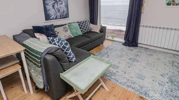 A living room with a sofa, a small table, a rug, and a view of the sea through a window. - Sea Snug (Berwick-upon-Tweed)