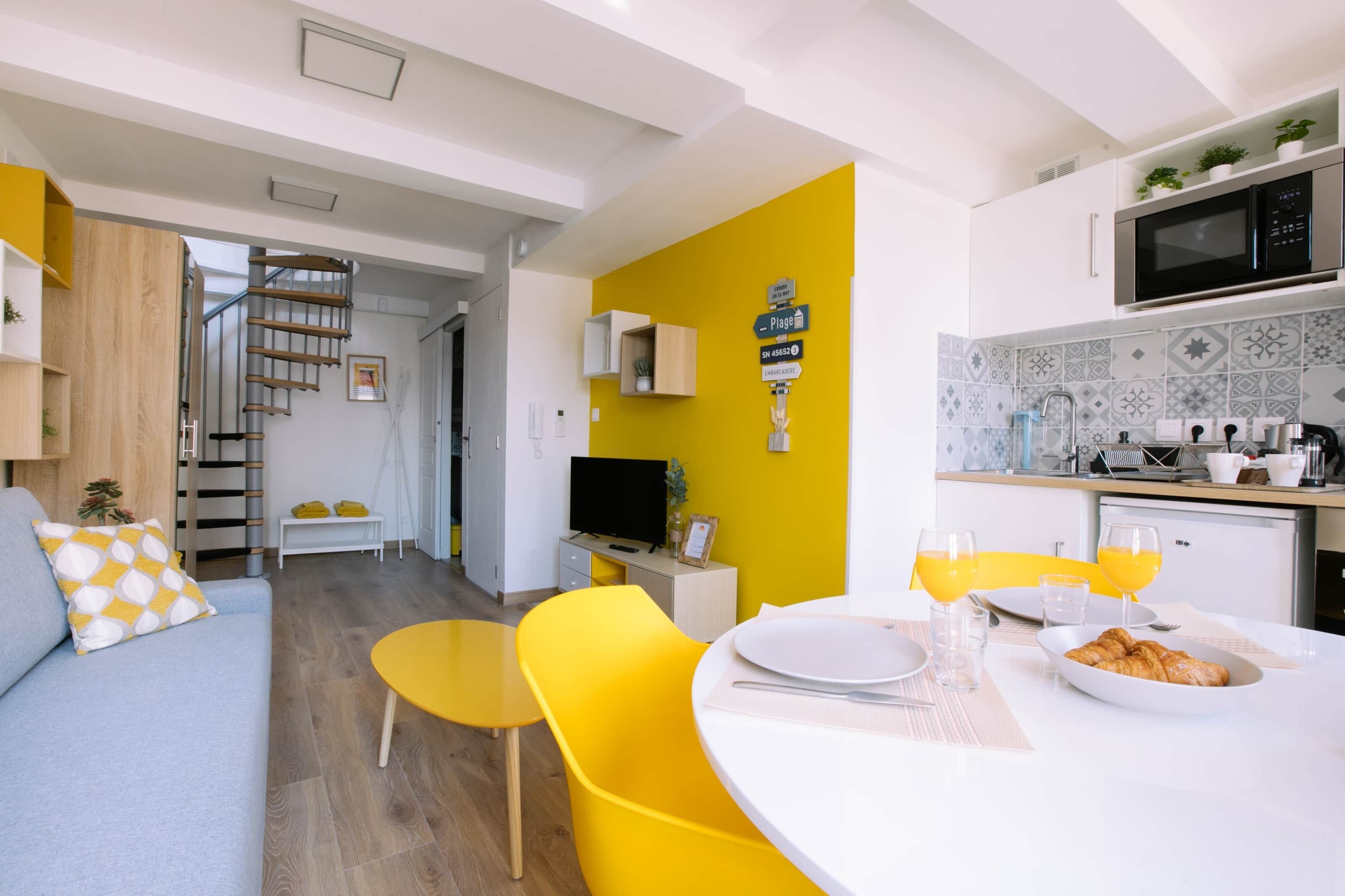 A modern living space with a staircase, a dining area with yellow chairs, and a kitchen with a microwave.