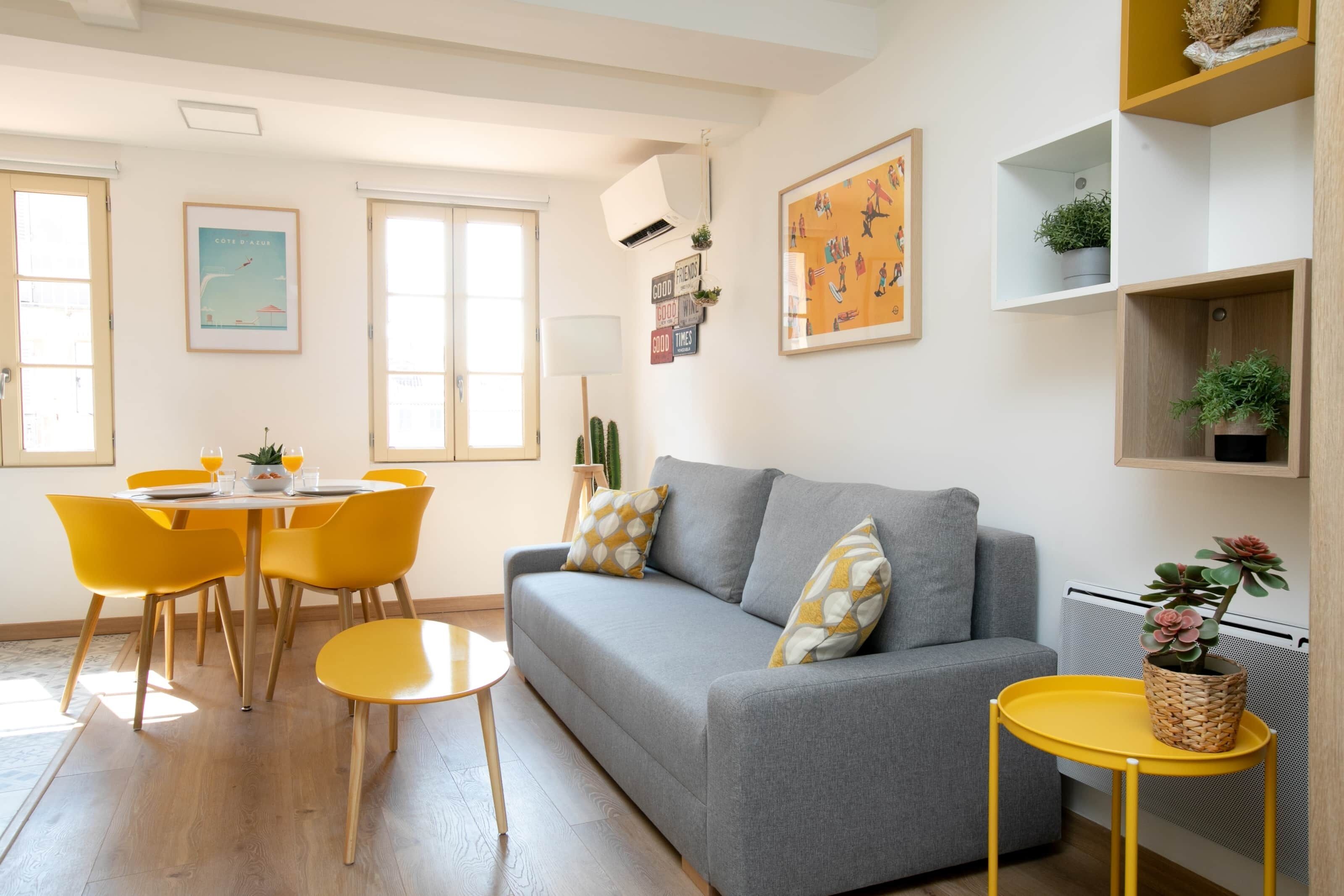 Ein modernes Wohnzimmer mit einem grauen Sofa, gelben Sesseln, einem kleinen runden Tisch und einem gelben Beistelltisch mit einer Pflanze.