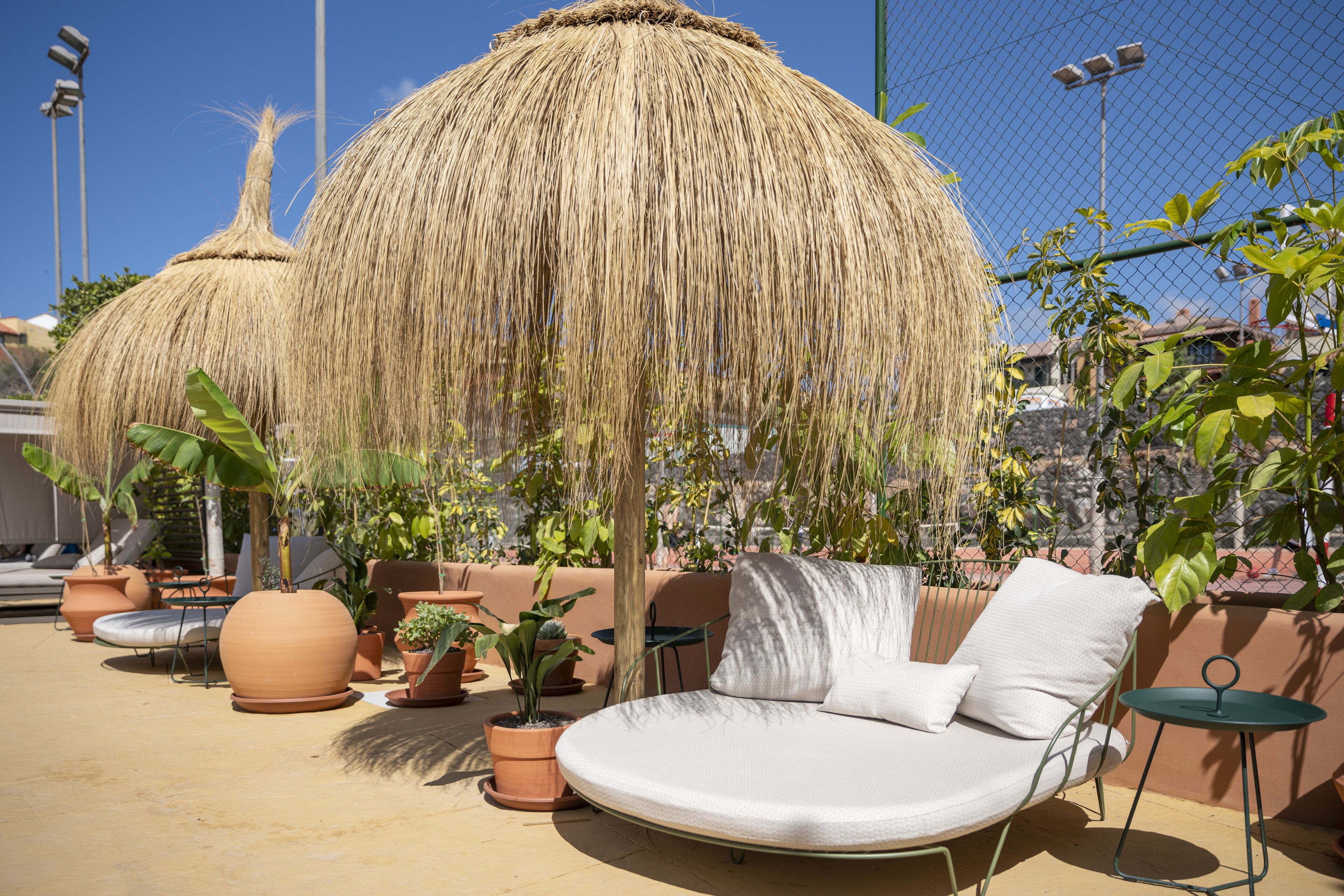 A sunlit outdoor seating area with a round cushioned chair, potted plants, and a thatched umbrella.