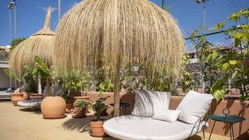 A sunlit outdoor seating area with a round cushioned chair, potted plants, and a thatched umbrella.