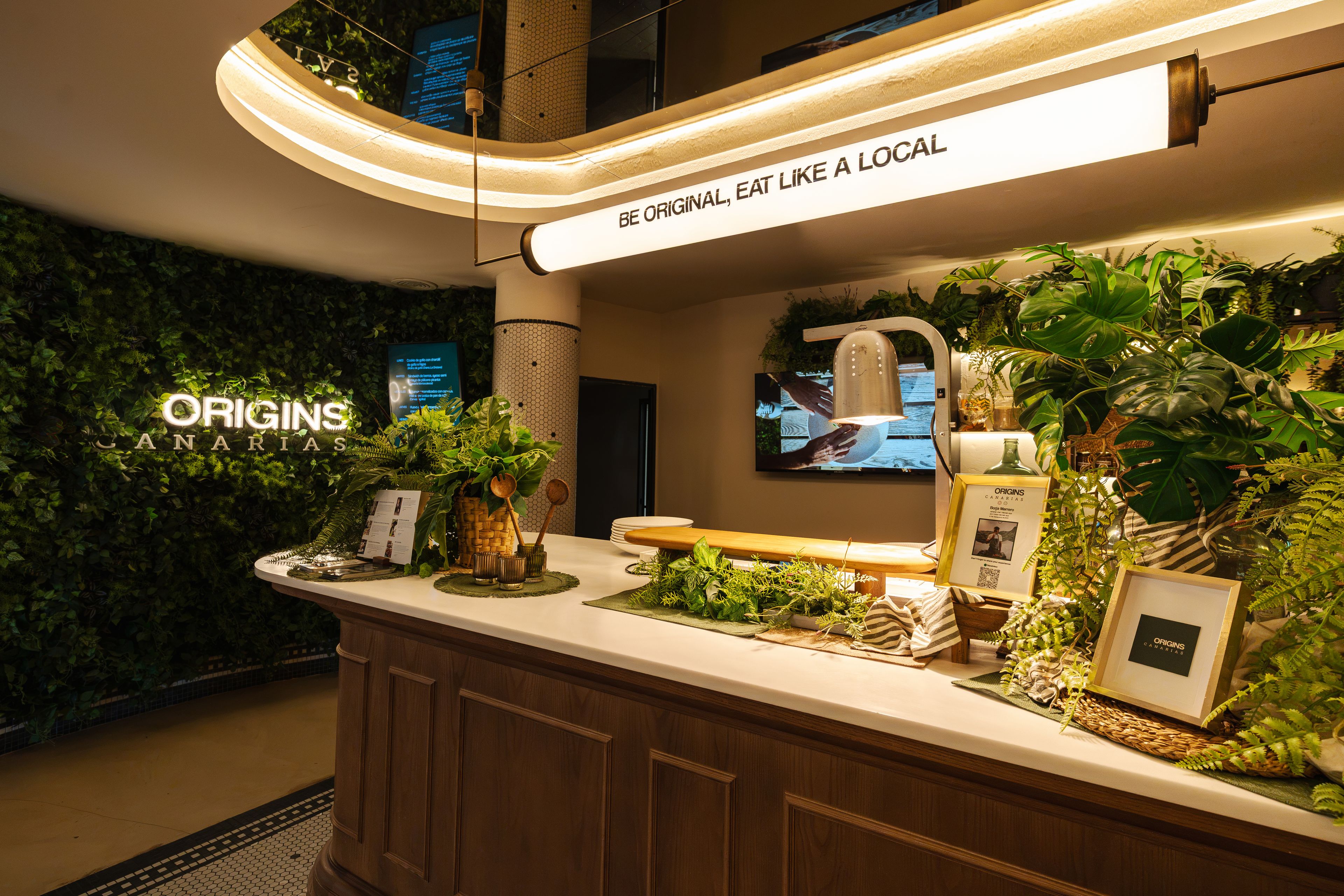 Een receptiebalie met een bord waarop 'ORIGINS CANARIAS' staat, en een slogan: 'WEES OUDENIJK, EET ALS EEN LOCAL'.